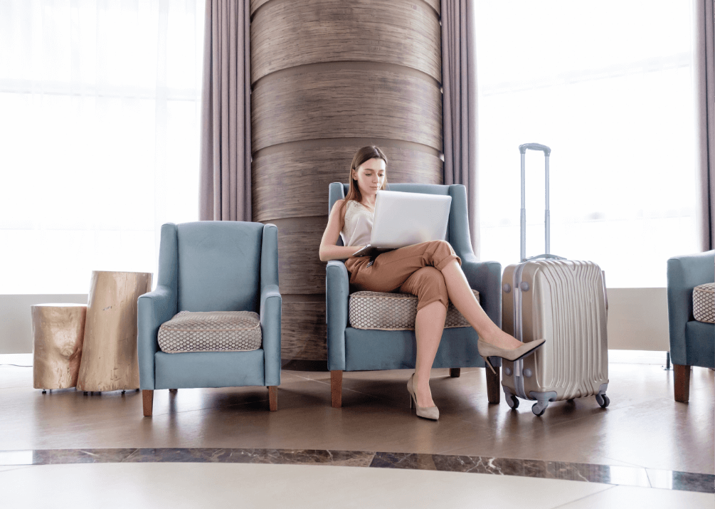 A woman sitting on a chair with a laptop on her knee and a suitcase beside her.